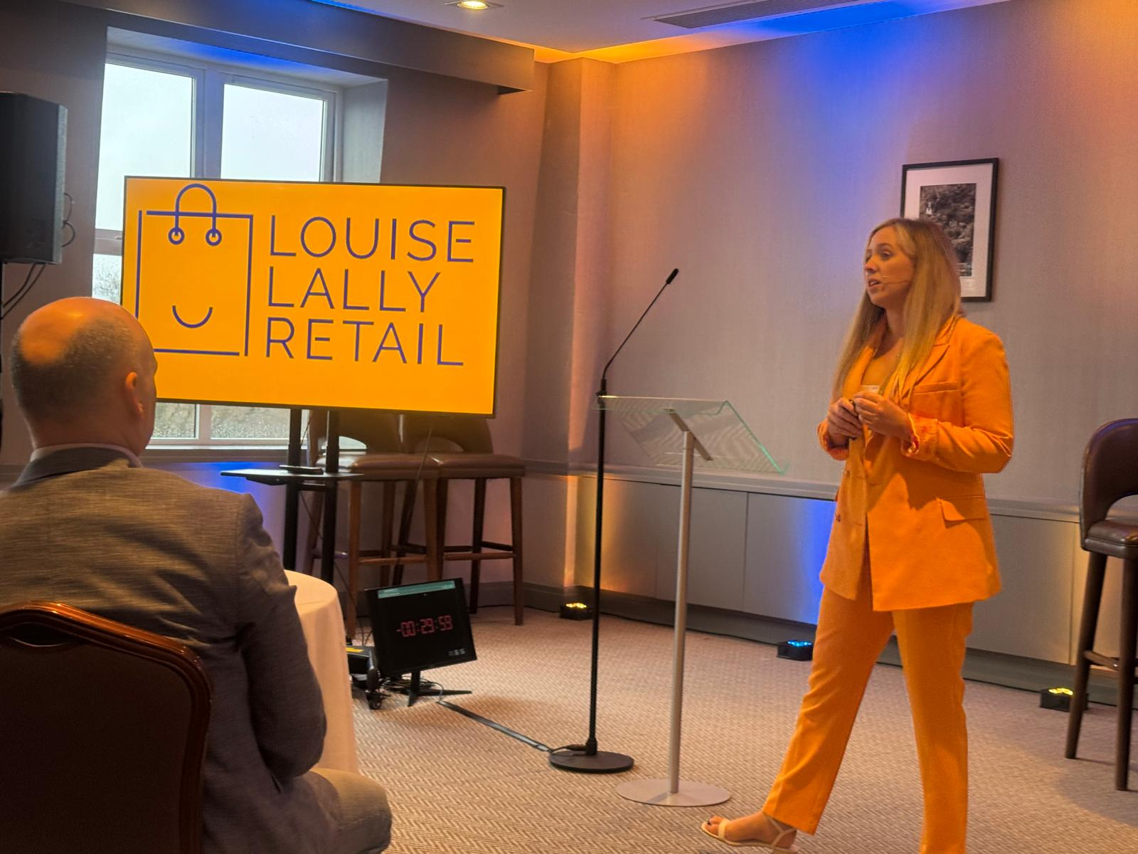 Louise Lally speaking at a retail industry event, wearing an orange suit in front of a Louise Lally Retail logo screen